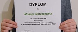 Zdjęcie przedstawia ucznia Miłosza Matyszczaka trzymającego w rękach dyplom za zdobycie trzeciej nagrody na Marcowym Konkursie Klarnetowym 2023 w Szczecinie.