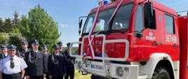 Samochód ratowniczo -gaśniczy pośród którego stoją strażacy w mundurach.