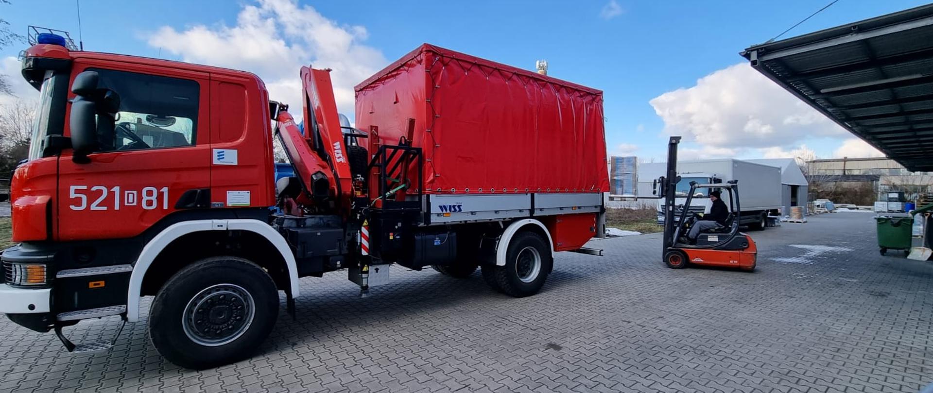 Na zdjęciu widać duży ciężarowy, transportowy samochód strażacki z oławskiej komendy straży pożarnej. Trwa załadunek ulotkami informacyjnymi o szczepieniach. Widać wózek widłowy ładujący kolejną partię ulotek na pakę samochodu.