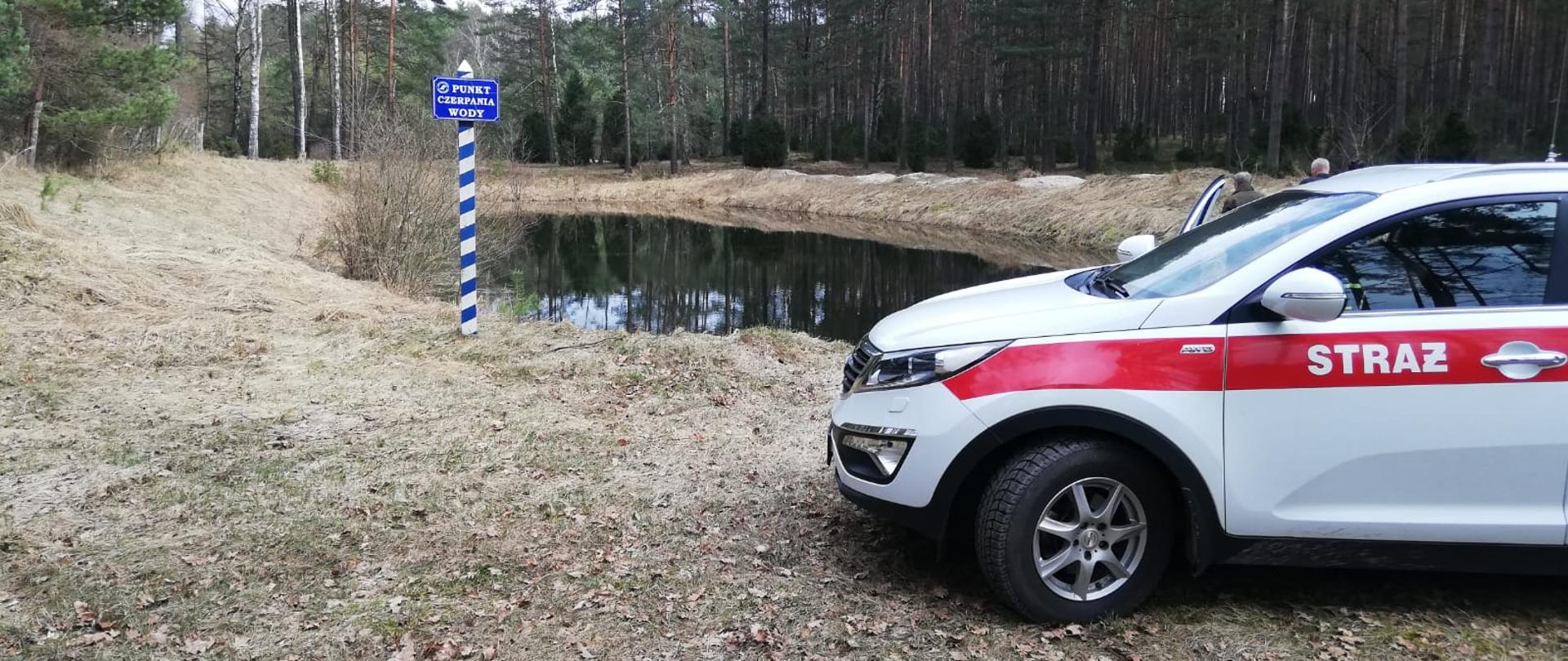 Na zdjęciu z prawej strony samochód operacyjny Komendy Powiatowej Państwowej Straży Pożarnej w Świeciu. Za samochodem jeden funkcjonariusz Komendy Powiatowej Państwowej Straży Pożarnej w Świeciu wraz z leśniczym lasów Państwowych podczas sprawdzania dostępności punktu czerpania wody na wypadek powstania pożaru w lesie.