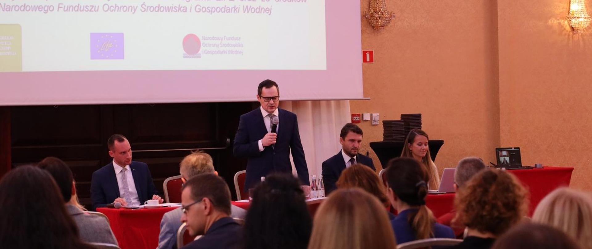 Widoczni ludzie siedzący za stołem, za nimi, ekran. Mężczyzna w garniturze stoi i mówi do mikrofonu. Z jego lewej strony siedzą kolejno mężczyzna w garniturze, a za nim kobieta spoglądająca w monitor komputera. Z lewej stron stającego mężczyzny siedzi mężczyzna w garniturze.