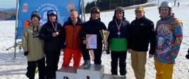 Zdjęcie przedstawia sześciu zawodników województwa podkarpackiego ubranych w stroje narciarskie oraz snowboardowe w asyście komendanta szkoły aspirantów w Krakowie. Wyróżnieni zawodnicy stoją za podium z zawieszonymi medalami, pucharem oraz dyplomem. W tle widoczny baner szkoły aspirantów PSP oraz stok narciarski 