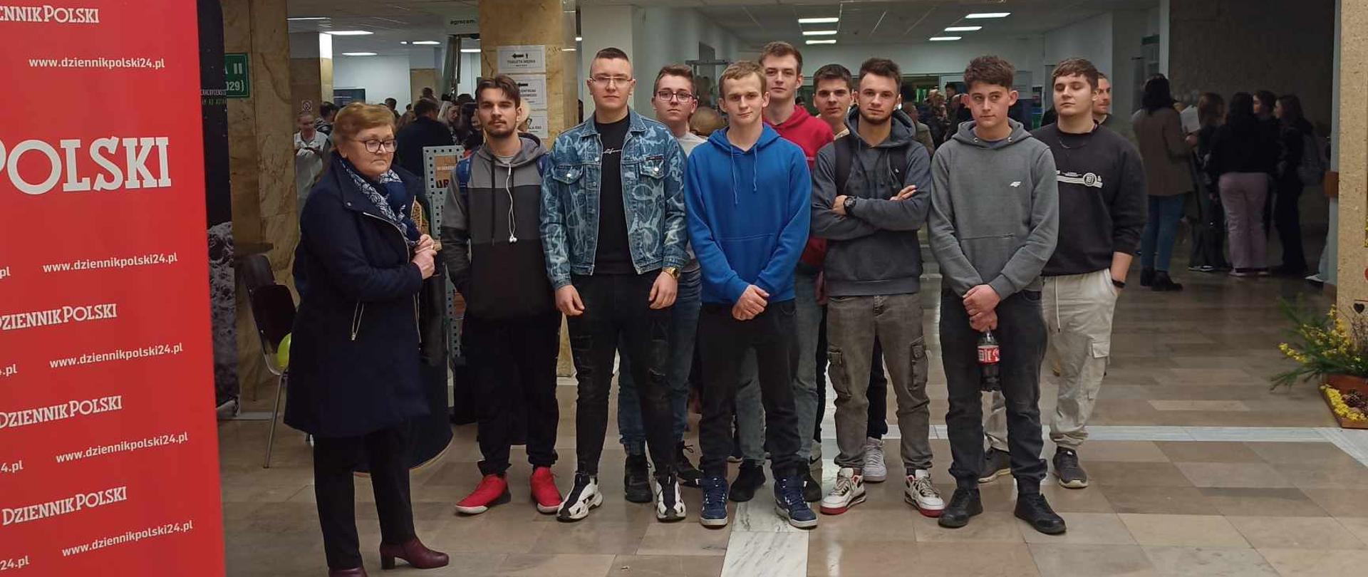 Grupa studentów i starsza kobieta pozują do wspólnego zdjęcia w przestronnym holu budynku. Na ścianie powyżej wisi trofeum w postaci głowy żubra