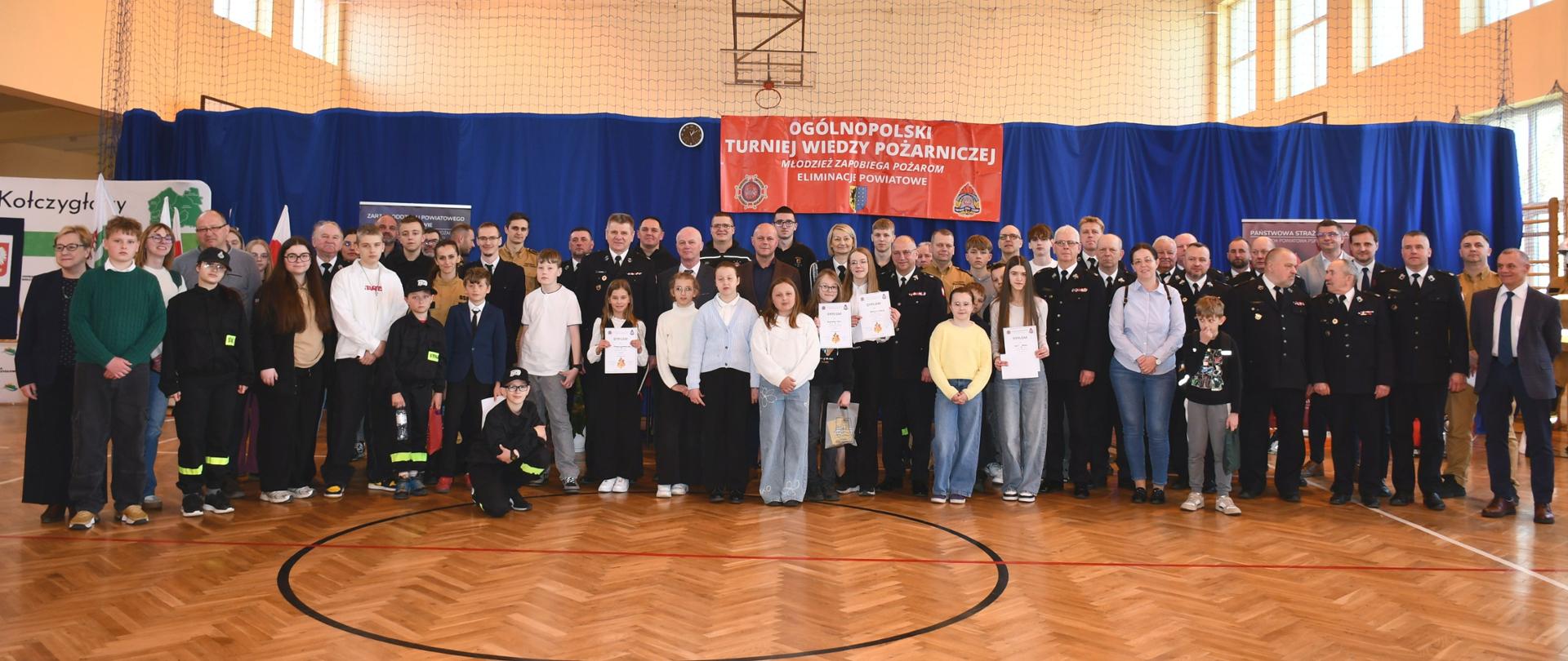 Na drewnianym parkiecie hali sportowej stoi liczna, wielopokoleniowa grupa uczestników i organizatorów. W pierwszym rzędzie widać dzieci i młodzież, z których kilkoro trzyma w rękach białe dyplomy. W kolejnych rzędach stoją dorośli – po bokach i w tle widocznych jest wielu strażaków w mundurach oraz osoby cywilne.
W tle, za grupą, na niebieskiej kurtynie wisi duży czerwony baner z białym napisem: „OGÓLNOPOLSKI TURNIEJ WIEDZY POŻARNICZEJ” oraz „ELIMINACJE POWIATOWE”. Po lewej stronie widać tablicę z nazwą „Kołczygłowy”. Nad głowami widać metalową konstrukcję dachu hali z siatką ochronną.