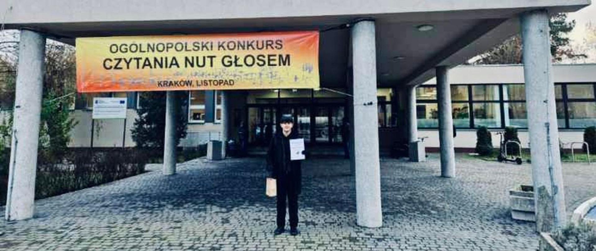 Na zdjęciu widać wejście do Państwowej Szkoły Muzycznej w Krakowie, nad którym wisi duży baner z napisem „Ogólnopolski Konkurs Czytania Nut Głosem – Kraków, listopad”. Przed wejściem, na brukowanym placu, stoi nasz uczeń Tobiasz Ganobis ubrany na ciemno, trzymająca w rękach dyplom i małą torbę. Budynek ma duże okna i kolumny podtrzymujące zadaszenie. W tle widać drzwi wejściowe oraz fragmenty zieleni po bokach.