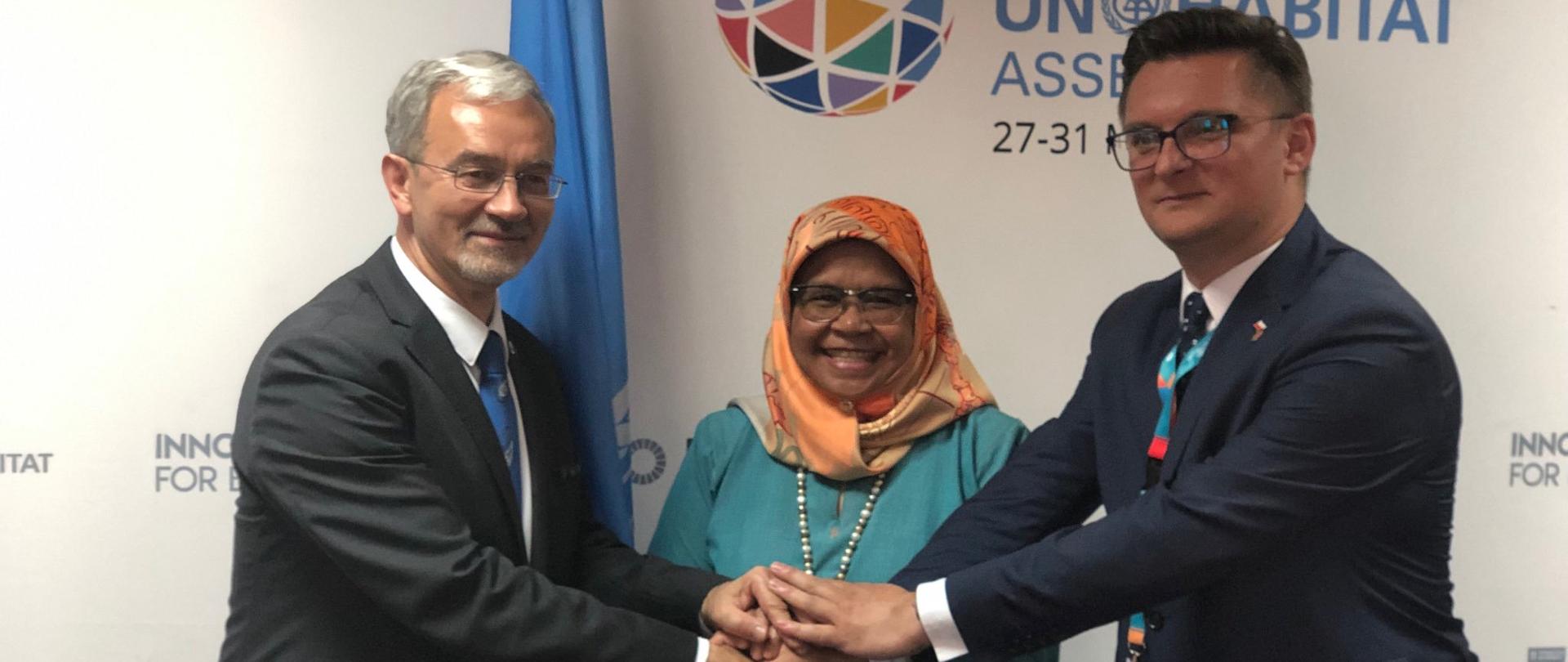 Na zdjęciu znajdują się trzy osoby przed ścianką promocyjną - minister Jerzy Kwieciński, Dyrektor Wykonawcza UN-HABITAT Maimunah Mohd Sharif i prezydent Katowic Marcin Krupa, trzymają się za ręce.