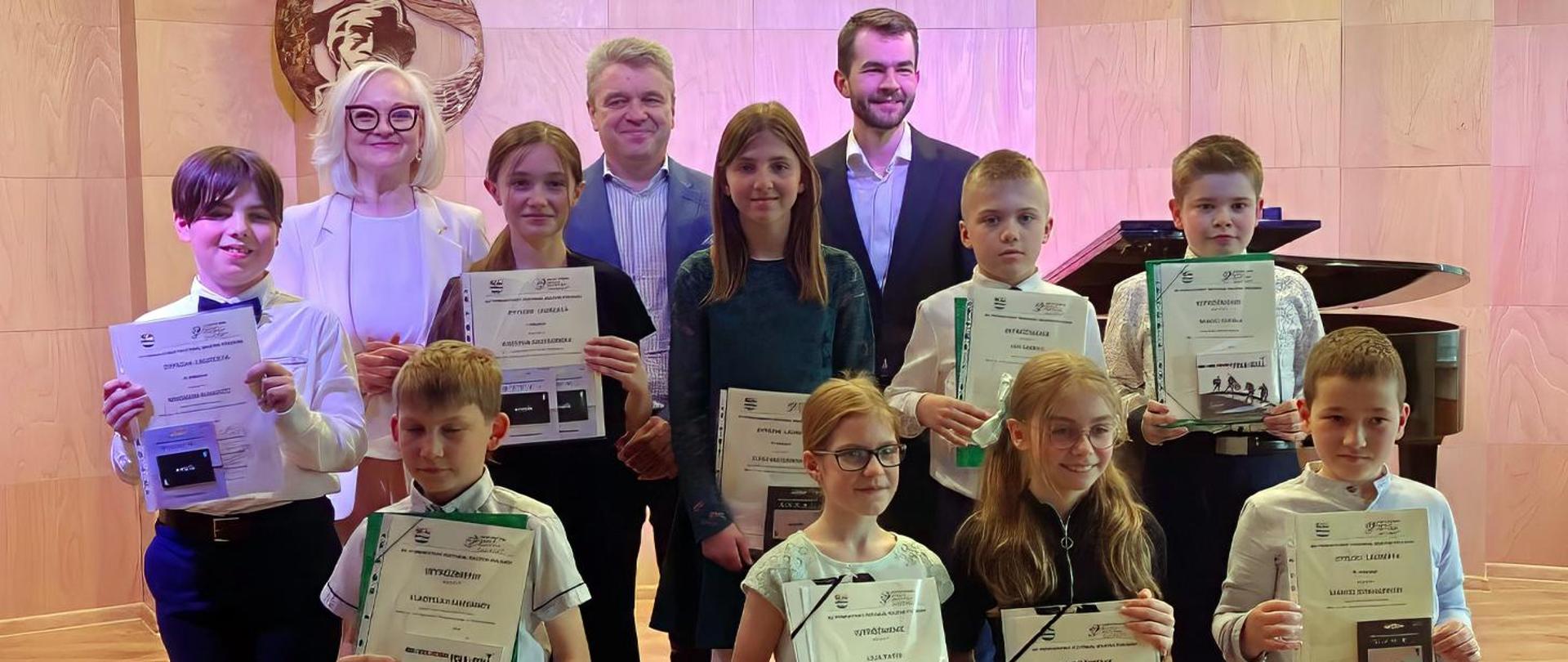 Na zdjęciu kilkunastoosobowa grupa dzieci i dorosłych stojąca na scenie, za nimi stoi fortepian a na ścianie wisi popiersie Fryderyka Chopina. Dzieci w rękach trzymają dyplomy.