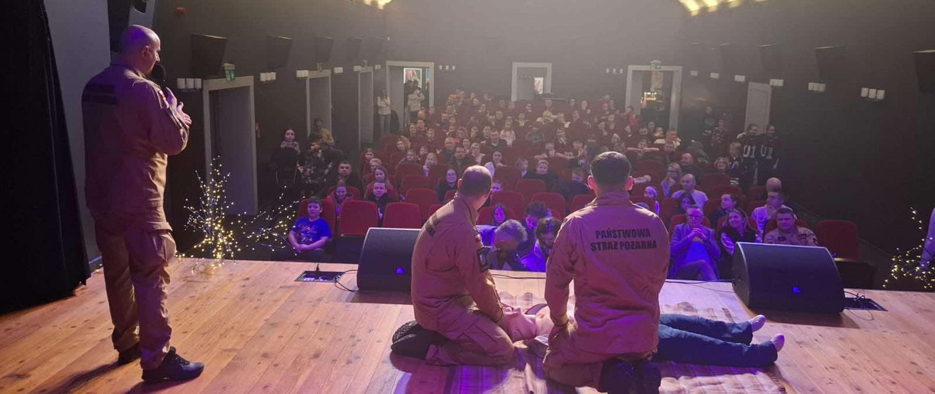 Na zdjęciu widać pokaz lub szkolenie z zakresu bezpieczeństwa publicznego, odbywające się na scenie w audytorium lub teatrze. Jedna osoba, ubrana w mundur, stoi po lewej stronie i prowadzi prezentację, trzymając mikrofon. Na środku sceny dwie inne osoby, również w mundurach z napisem "PAŃSTWOWA STRAŻ POŻARNA" na plecach jednej z nich, klęczą obok osoby leżącej na podłodze, demonstrując techniki pierwszej pomocy lub resuscytacji. Publiczność siedzi w rzędach czerwonych krzeseł, obserwując pokaz. Scena jest oświetlona reflektorami, a po lewej stronie sceny znajdują się ozdobne światełka.