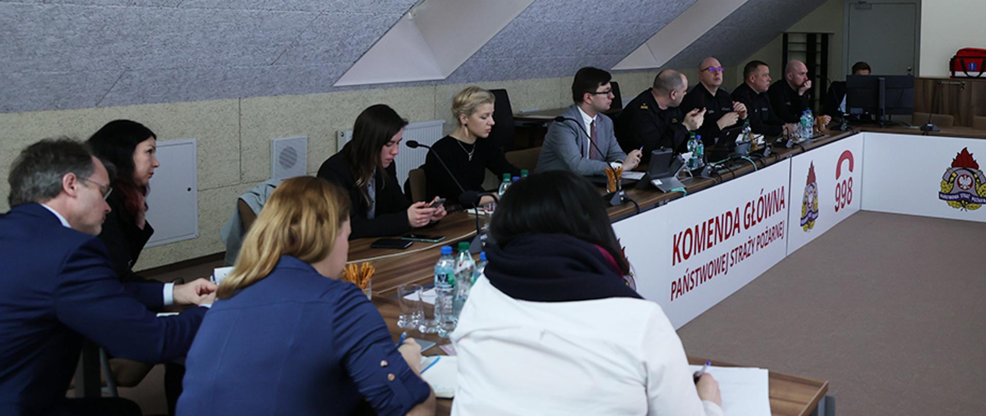 Pięć kobiet i siedmiu mężczyzn siedzący przy stole ułożonym "w podkowę" Widoczny napis Komenda Główna Państwowej Straży Pożarnej