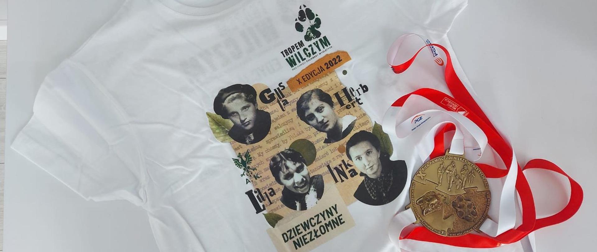 Zdjęcie przedstawia białą koszulkę, na której znajdują się twarze czterech kobiet wraz z ich pseudonimami a pod spodem napis: "Dziewczyny niezłomne". Na koszulce leży również medal z biało czerwoną smyczką. Na medalu widnieje postać wilka odbita łapa oraz osoby biegnące. 