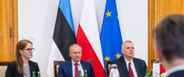 Zdjęcie przedstawia polską delegację siedzącą przy stole: ministra Tomasza Siemoniaka (po prawej), Mariusza Tomaszewskiego, dyrektora Departamentu Koordynacji Służb Specjalnych KPRM (w środku) oraz Agnieszkę Pacek, zastępcę dyrektora DKSS KPRM (po lewej). Za delegacją stoją trzy flagi w konfiguracji od prawej: UE (niebieska z żółtymi gwiazdkami), Polski (biało-czerwona), Estonii (niebiesko-czarno-biała). Po prawej i lewej stronie zdjęcia widać fragmenty drewnianych drzwi. Na stole: termosy z kawą, filiżanki, szklanki, butelki z wodą, talerzyki z przekąskami, kompozycja kwiatowa oraz biało-czerwony proporczyk. W prawym dolnym rogi widać siedzącego tyłem członka estońskiej delegacji.