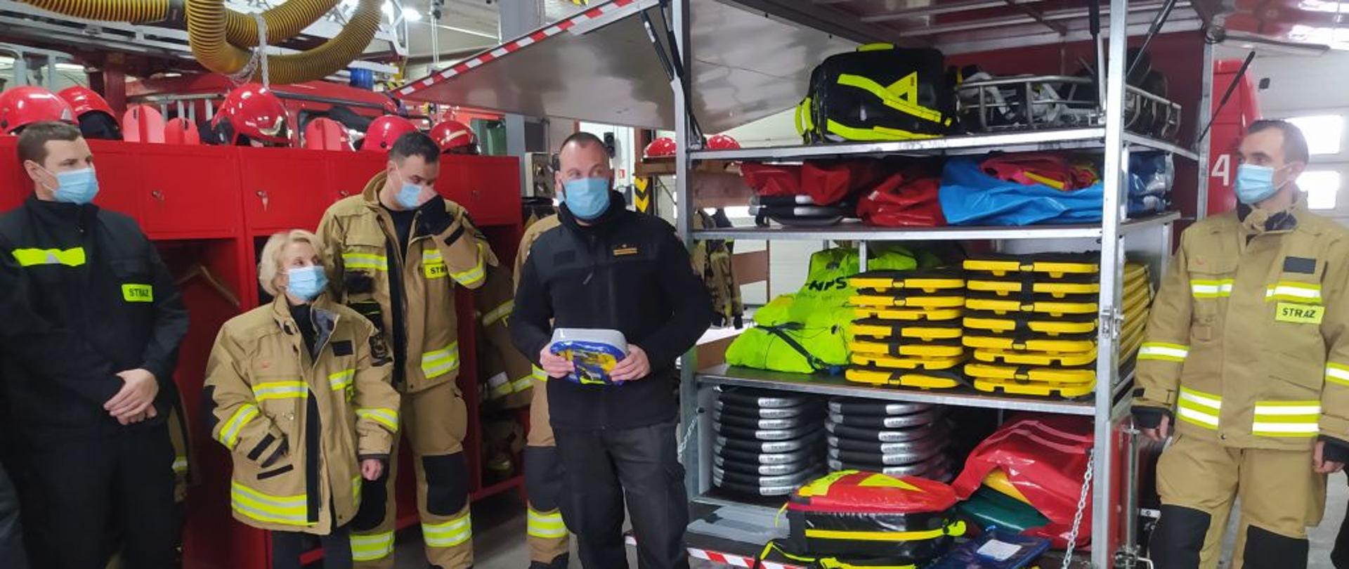 W garażu po bokach na tle czerwonych szafek stoją strażacy w żółtych i czarnych mundurach. Po środku wielka skrzynia z otwartymi drzwiami wewnątrz widać żółte czerwone rzeczy.