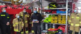 W garażu po bokach na tle czerwonych szafek stoją strażacy w żółtych i czarnych mundurach. Po środku wielka skrzynia z otwartymi drzwiami wewnątrz widać żółte czerwone rzeczy.