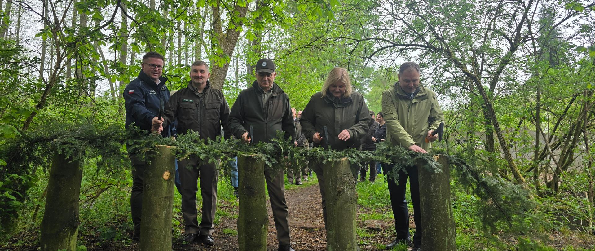 Pięć osób stoi na leśnej ścieżce wśród gęstej zieleni i bierze udział w symbolicznym otwarciu lub inauguracji. Każda osoba trzyma młotek i stoi przy drewnianych słupkach połączonych świerkowymi gałęziami. Uczestnicy są ubrani w kurtki terenowe i odzież leśną. W tle widać kolejne osoby obserwujące wydarzenie oraz drzewa z wiosennymi liśćmi.
