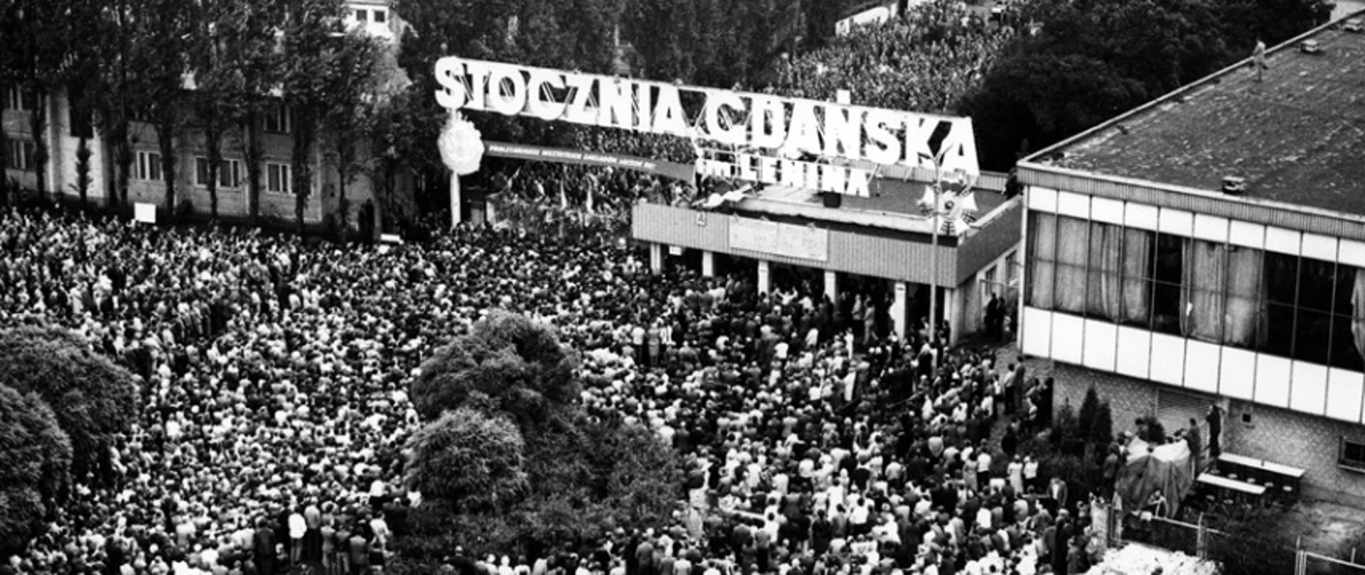 August strike at Lenin's Shipyard in Gdansk. Top view of Gate No. 2 and the citizens and shipyard workers gathered below it. Above the gate, there is a banner with the slogan "PROLETARIANS OF ALL FACTORIES UNITE!", and on the roof of the guardhouse there are boards with 21 demands of MKS.