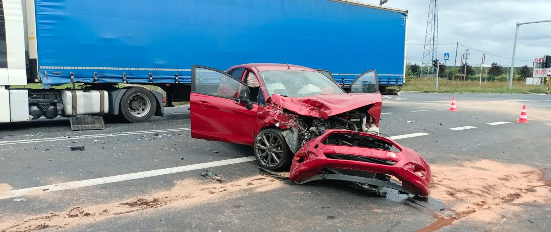 Widać uszkodzony czerwony samochód osobowy na środku, otwarte drzwi, za nim niebieska naczepa samochodu ciężarowego