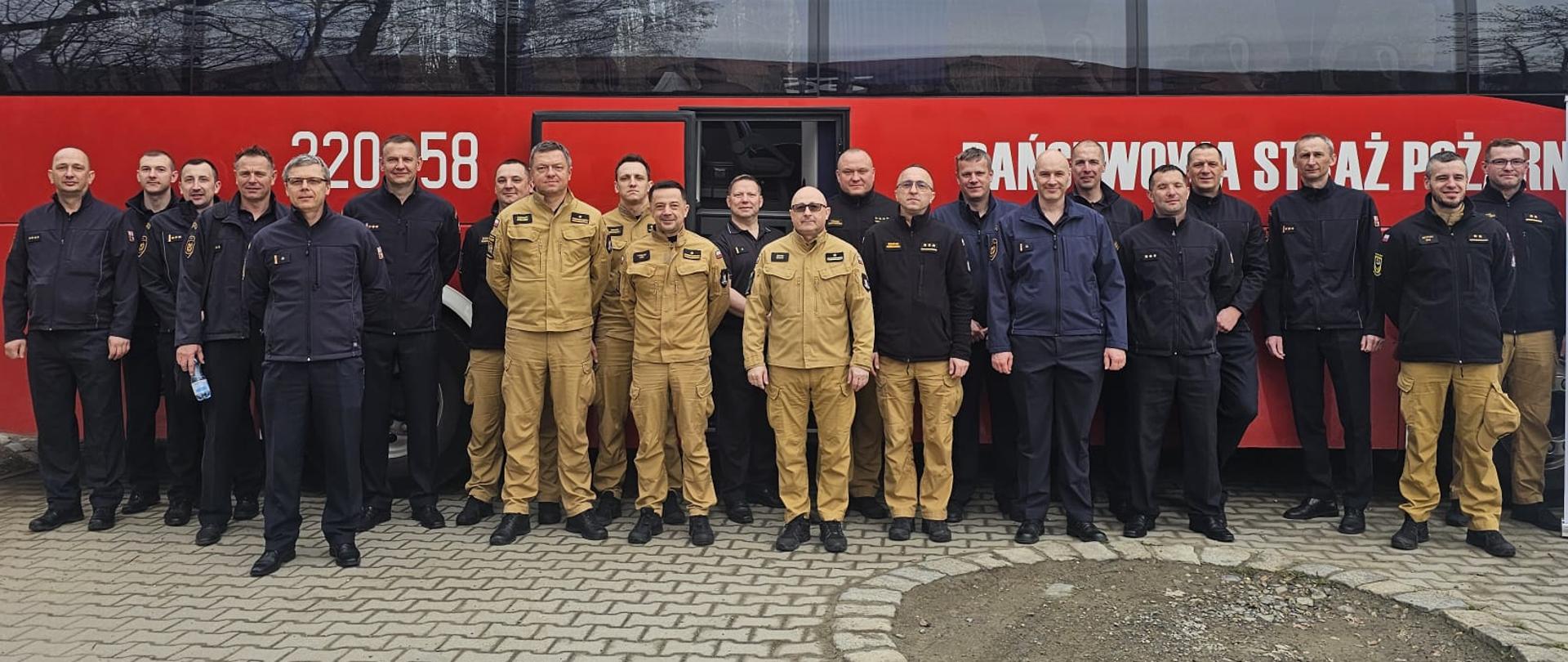 Zdjęcie grupowe uczestników spotkania na zewnątrz, przed dużym, czerwonym autokarem strażackim z napisem „PAŃSTWOWA STRAŻ POŻARNA”. Około dwudziestu mężczyzn w mundurach (polskich i czeskich) stoi w jednym rzędzie na wybrukowanym placu. Po prawej stronie widoczny jest biały roll-up z informacjami o projekcie Interreg Czechy – Polska, zawierający logotypy Unii Europejskiej oraz herb województwa opolskiego. W tle widać bezlistne drzewa, co sugeruje wczesną wiosnę lub jesień.