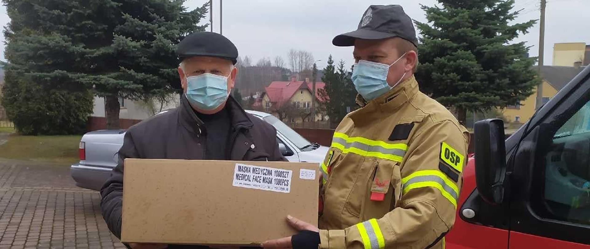Ochotnik przekazujący maseczki proboszczowi parafii na tle samochodu pożarniczego i samochodu osobowego.