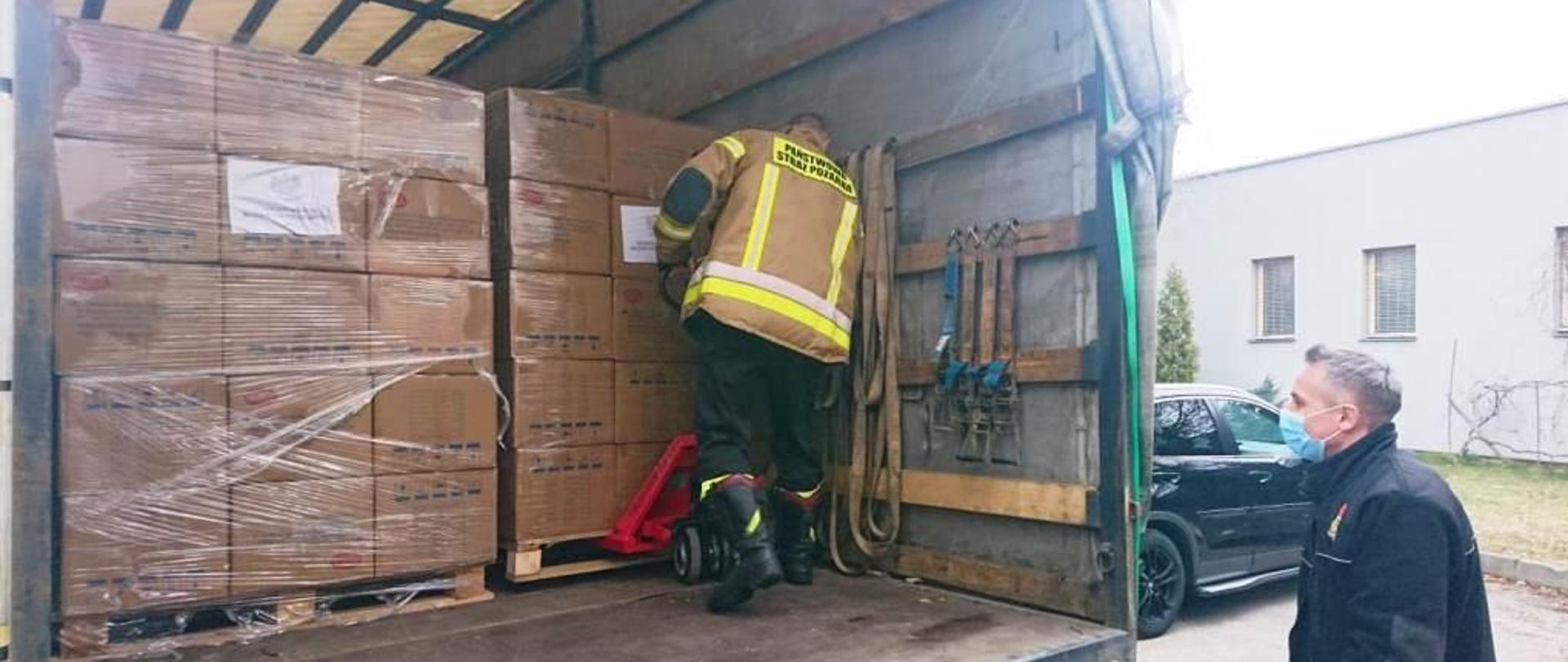 Zdjęcie przedstawia samochód ciężarowy na którym znajdują się opakowania z maseczkami chirurgicznymi. Przy opakowaniach znajduje się jeden ze strażaków, który przy pomocy wózka do transportu palet próbuje przewieźć jedną z palet jak najbliżej tylnej krawędzi pojazdu. Przy pojeździe stoi drugi strażak, który obserwuje transport palety. 
