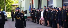 Oficer straży pożarnej maszeruje oddając honory przed pododdziałem funkcjonariuszy i pracowników cywilnych - łącznie kilkadziesiąt osób. Pododdział na tle budynku strażnicy. 