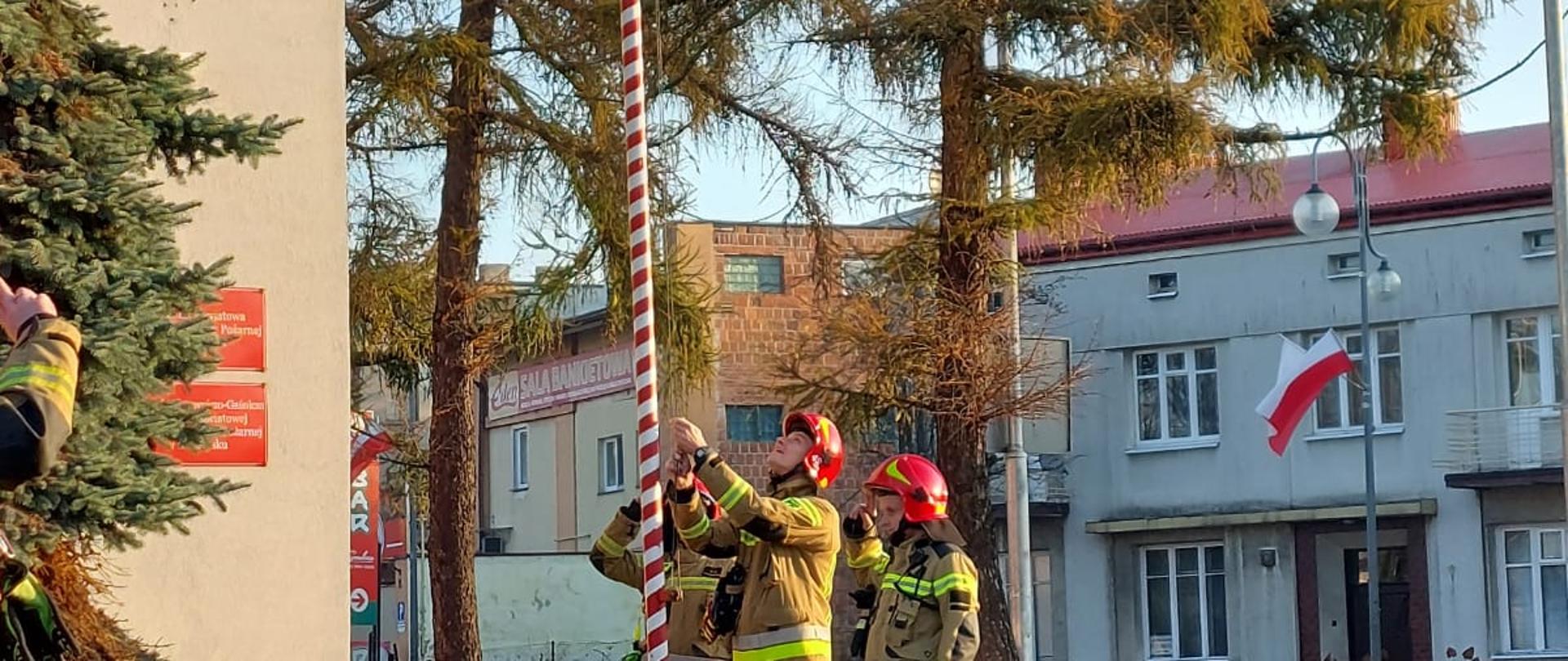 Trzech strażaków w ubraniach bojowych w czerwonych hełmach na głowie stojących obok masztu. Jeden z nich wciąga flagę Polski. W tle budynek komendy