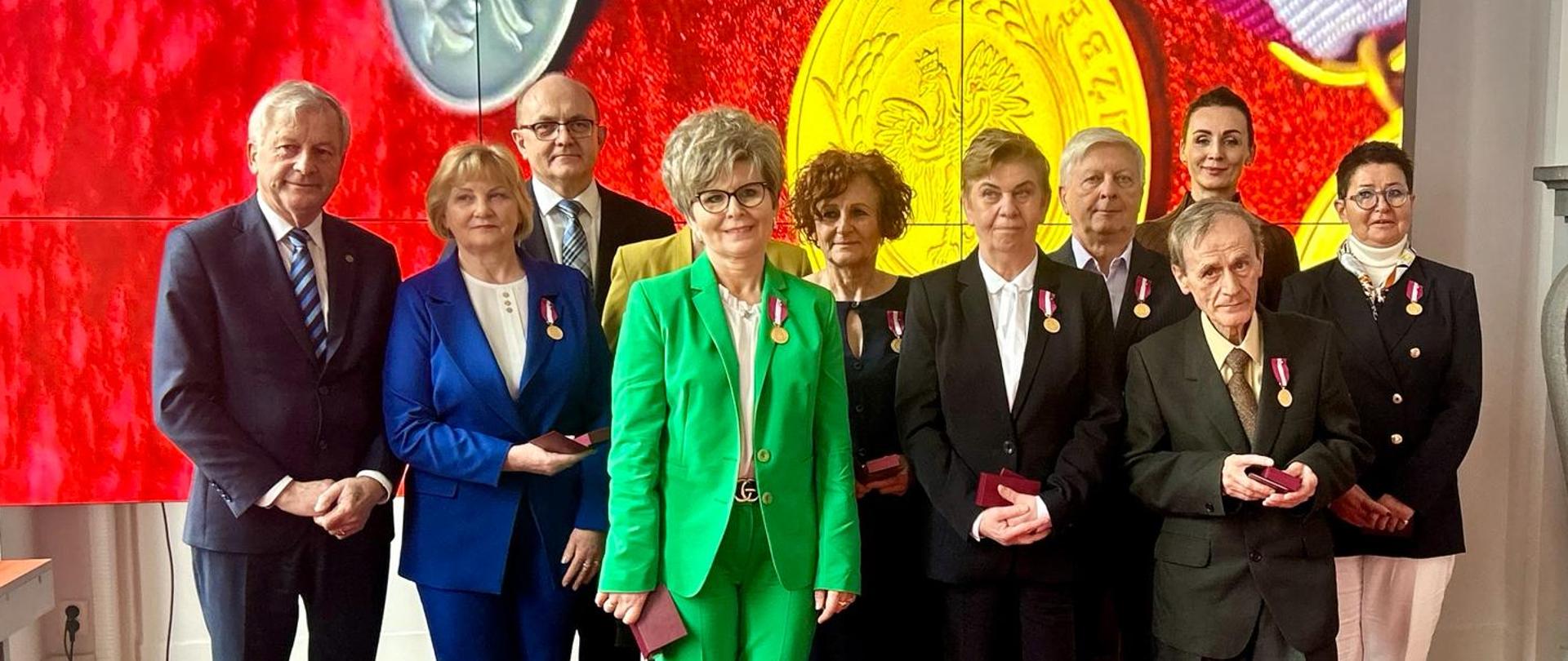 Grupa osób z medalami przypiętymi przy klapie stoi na tle obrazu w dużej sali urzędu wojewódzkiego