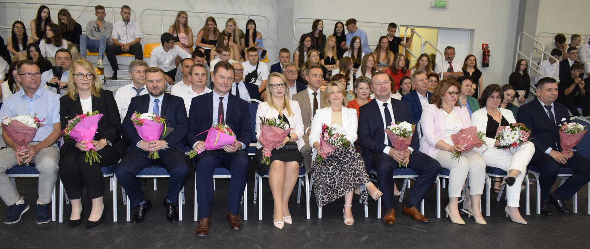 Grupa ludzi siedząca na krzesłach w wielkiej sali. Ubrani są na elegancko. W dłoniach trzymają bukiety kwiatów.