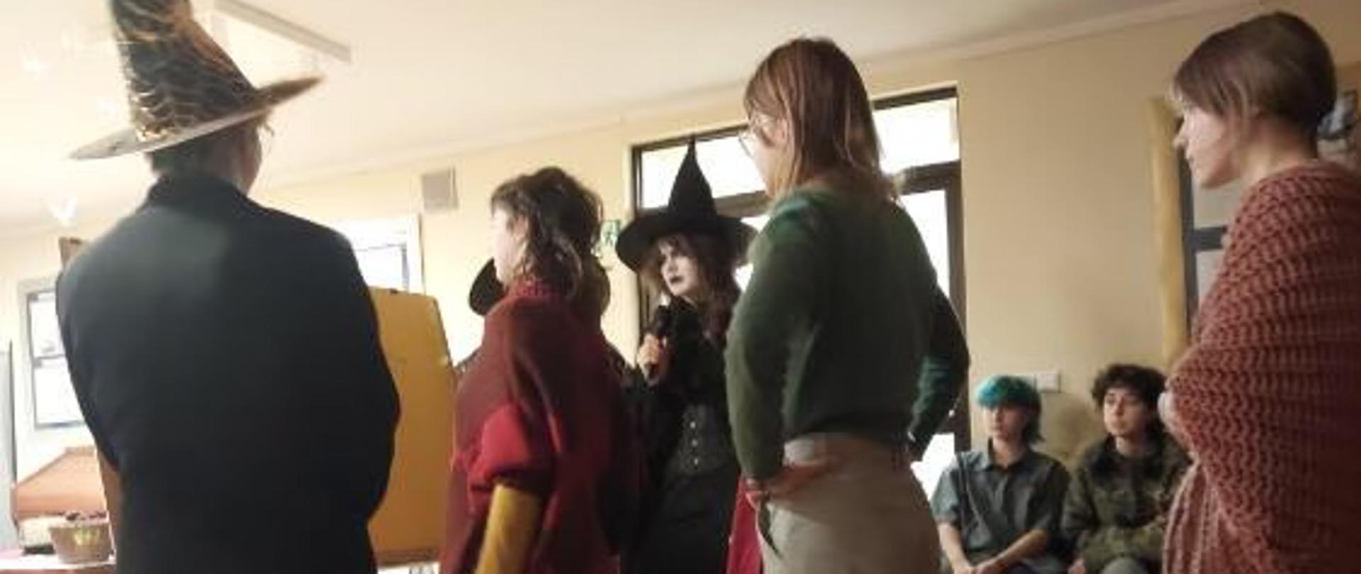 Zdjęcie z wieczoru andrzejkowego. Na sali spotkań zebrana młodzież. Część z nich przebrana w stroje czarownic - to osoby prowadzące imprezę. Część jest uczestnikami zabawy i wróżb andrzejkowych. Na zdjęciu przeprowadzany konkurs przy małej tablicy na sztaludze.