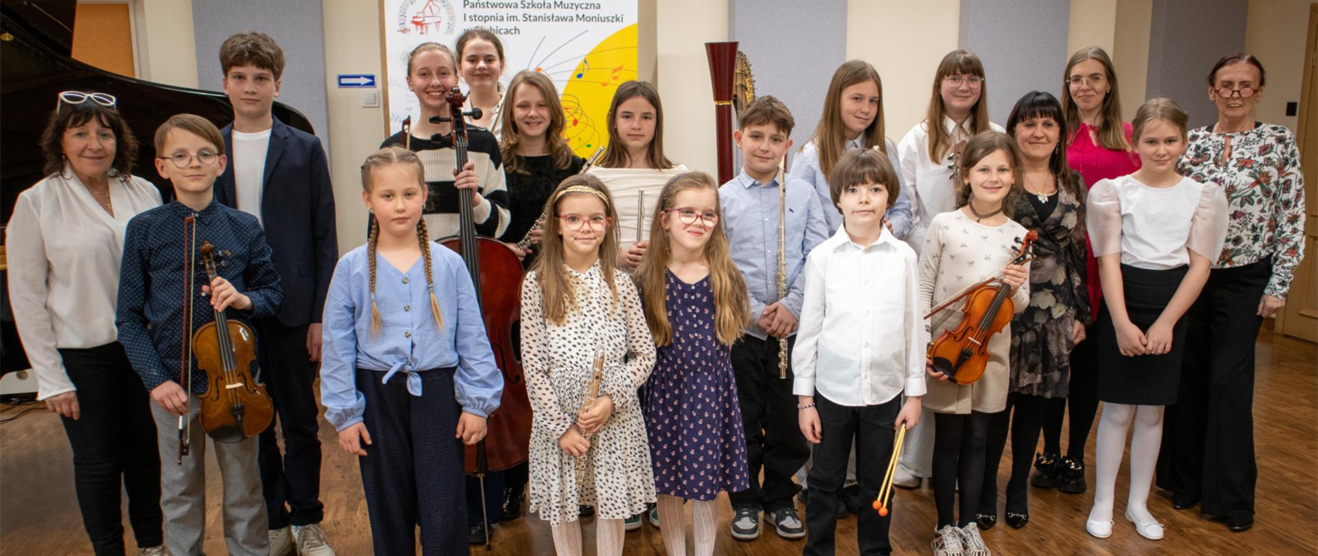 Grupa kilkunastu osób, nauczycieli i uczniów stoi w auli szkolnej na tle banera szkolnego.