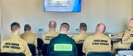 Grupa strażaków w beżowych mundurach PSP siedzi na krzesłach ustawionych w kierunku telewizora, na którym wyświetlana jest prezentacja. Wśród uczestników znajduje się także jedna osoba w ciemnym stroju z dużym napisem „STRAŻ” na plecach. Wszyscy są skupieni na treści slajdu dotyczącego zasad związanych z ochroną danych osobowych. 