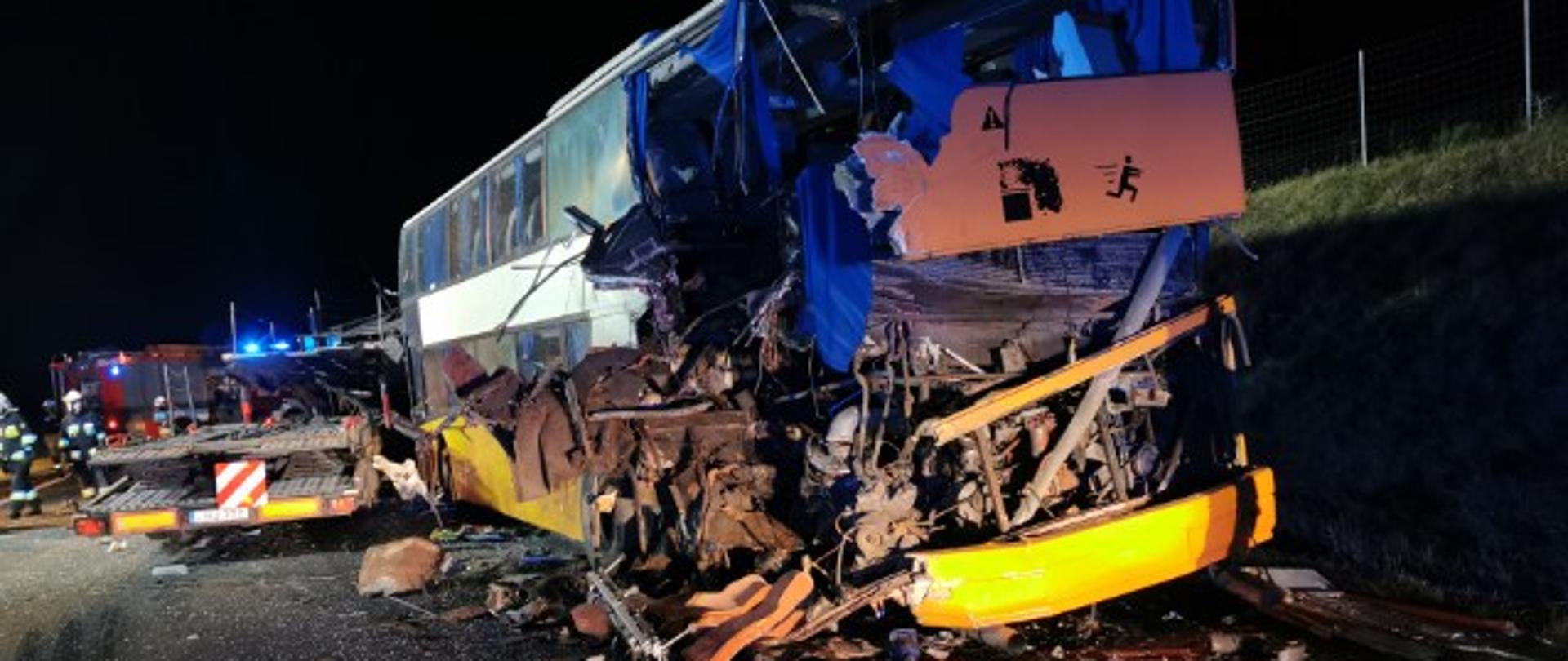 Zdjęcie przedstawia stojący na kołach dwupoziomowy autobus z rozbitym tyłem i częścią boku. Wokół autobusu leżą jego fragmenty. Za autobusem ustawiony jest tyłem samochód-laweta pomocy drogowej. W tle widać pracujących w ubraniach specjalnych strażaków z terenu powiatu zambrowskiego. Zdjęcie wykonano na drodze krajowej nr 8 na wysokości miejscowości Wiśniówek-Wertyce, gm. Kołaki Kościelne w pogodną noc. 