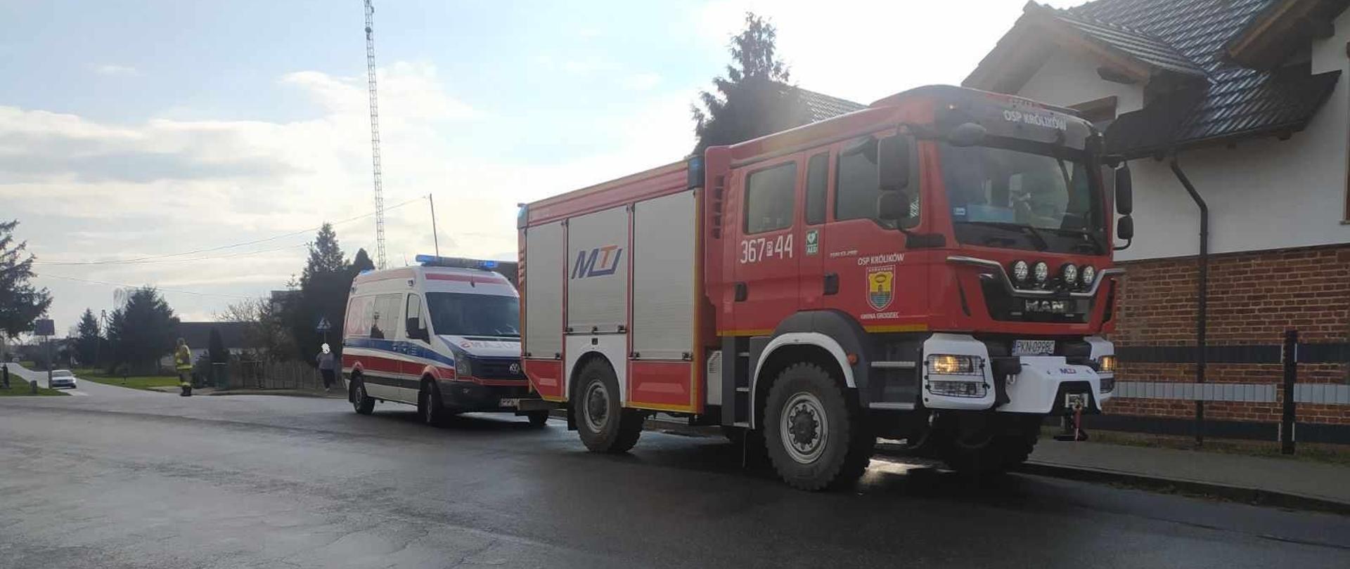 Zdjęcie przedstawia samochód strażacki oraz karetkę pogotowia ratunkowego, które stoją zaparkowane na jezdni przed budynkiem mieszkalnym jednorodzinnym