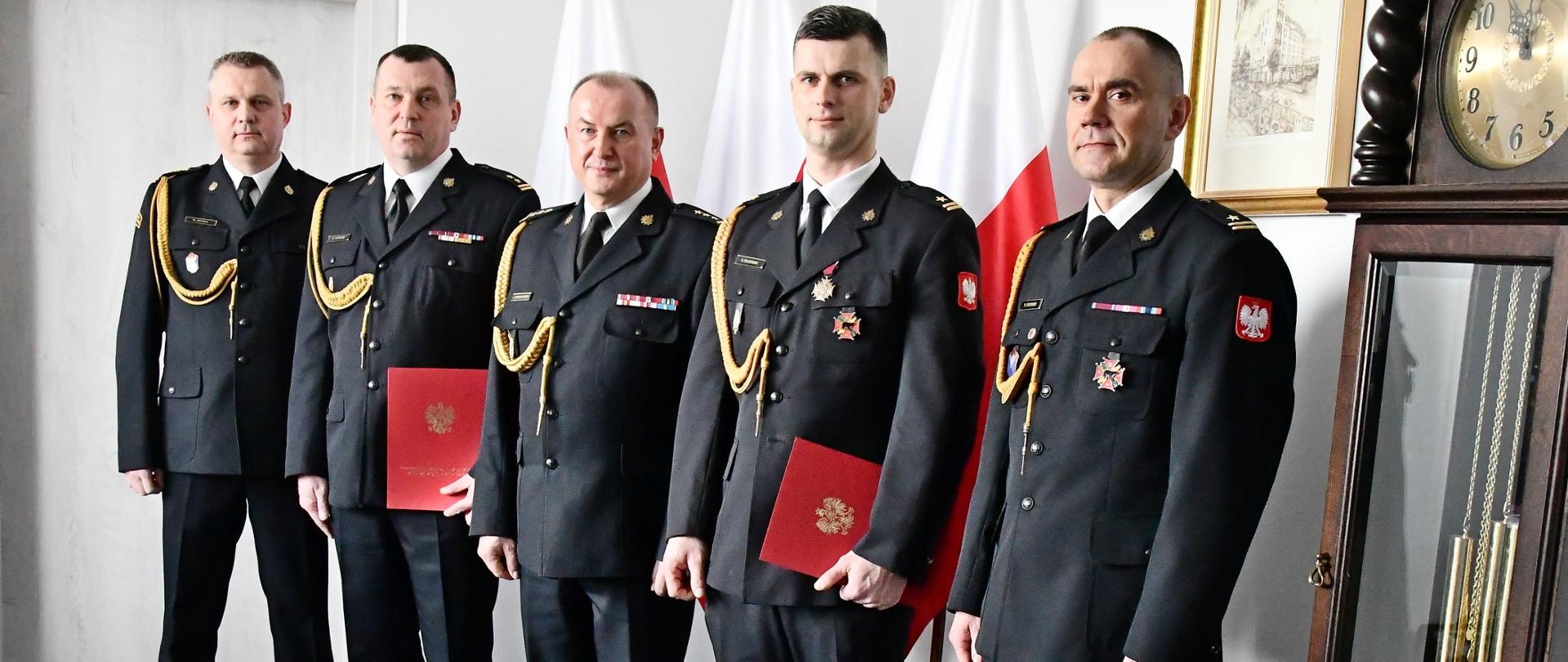 Pięciu strażaków w mundurach wyjściowych ze sznurem stoi obok siebie drugi i czwarty strażak trzyma teczkę za nimi są trzy flagi Polski na ścianie wisi obraz obok stoi zegar.