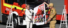 Zdjęcie przedstawia Minister Klimatu i Środowiska i Komendanta Głównego PSP prezentujących plakat promujący akcję "Bitwa o Remizy" na tle samochodów strażackich