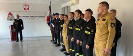 Uroczysta zbiórka z okazji ślubowania strażaka. Strażacy stoją w rzędzie w umundurowaniu. 