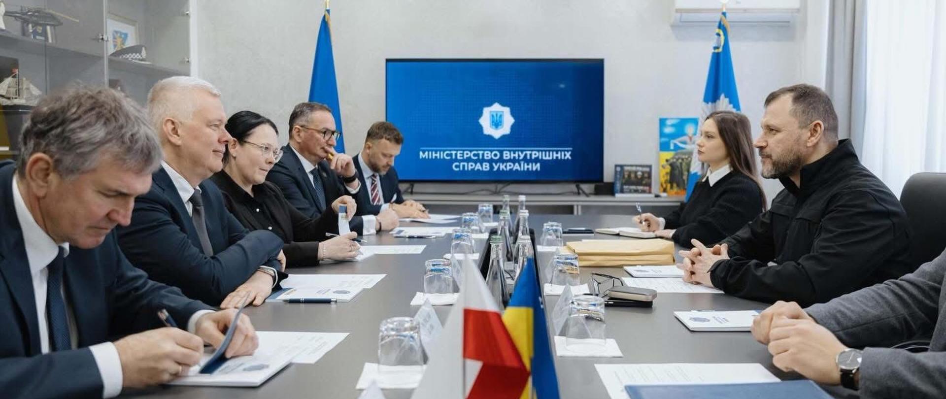 Na zdjęciu widać duży stół. Po lewej stronie stołu siedzi pięć osób. Na pierwszym planie po lewej siedzi minister Radosław Kujawa, jako drugi po lewej siedzi minister Tomasz Siemoniak. Po prawej stronie stołu siedzą trzy osoby - w środku siedzi szef MSW Ukrainy Ihor Klymenko. Na stole znajdują się teczki, notesy, dwa proporczyki z flagami polską i ukraińską, butelki z wodą, szklanki. W tle stołu znajduje się telewizor oraz dwie flagi. Po lewej stronie pokoju widać szklaną gablotę. 
