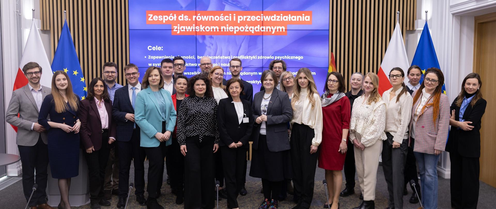 Zdjęcie zbiorowe - pod wbudowanym w ścianę z cienkich pionowych listewek wielkim ekranem z napisem Zespół ds. równości i przeciwdziałania zjawiskom niepożądanym w środowisku akademickim stoi kilkadziesiąt osób, pośrodku grupy wiceminister Zioło-Pużuk w szarej marynarce.