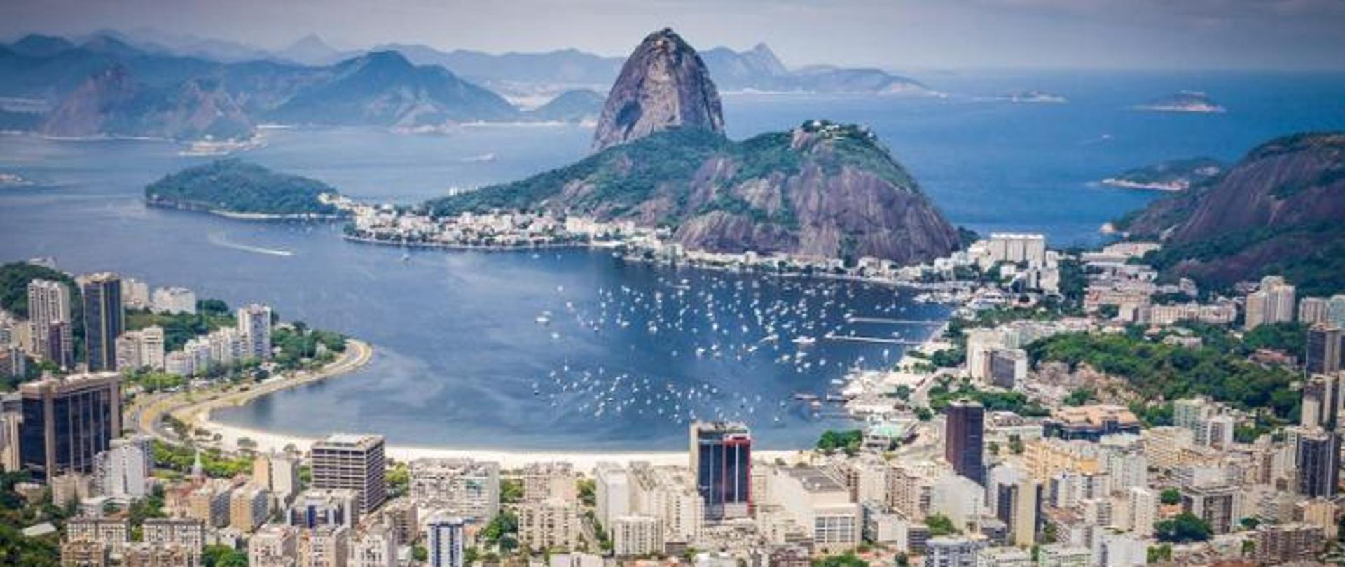 Panorama miasta Rio de Janeiro w Brazylii – na pierwszym planie widać nowoczesne budynki i plażę, a w tle charakterystyczna góra - Głowa Cukru otoczona zatoką z licznymi białymi łodziami. W tle rozciąga się ocean oraz wyspy na horyzoncie.