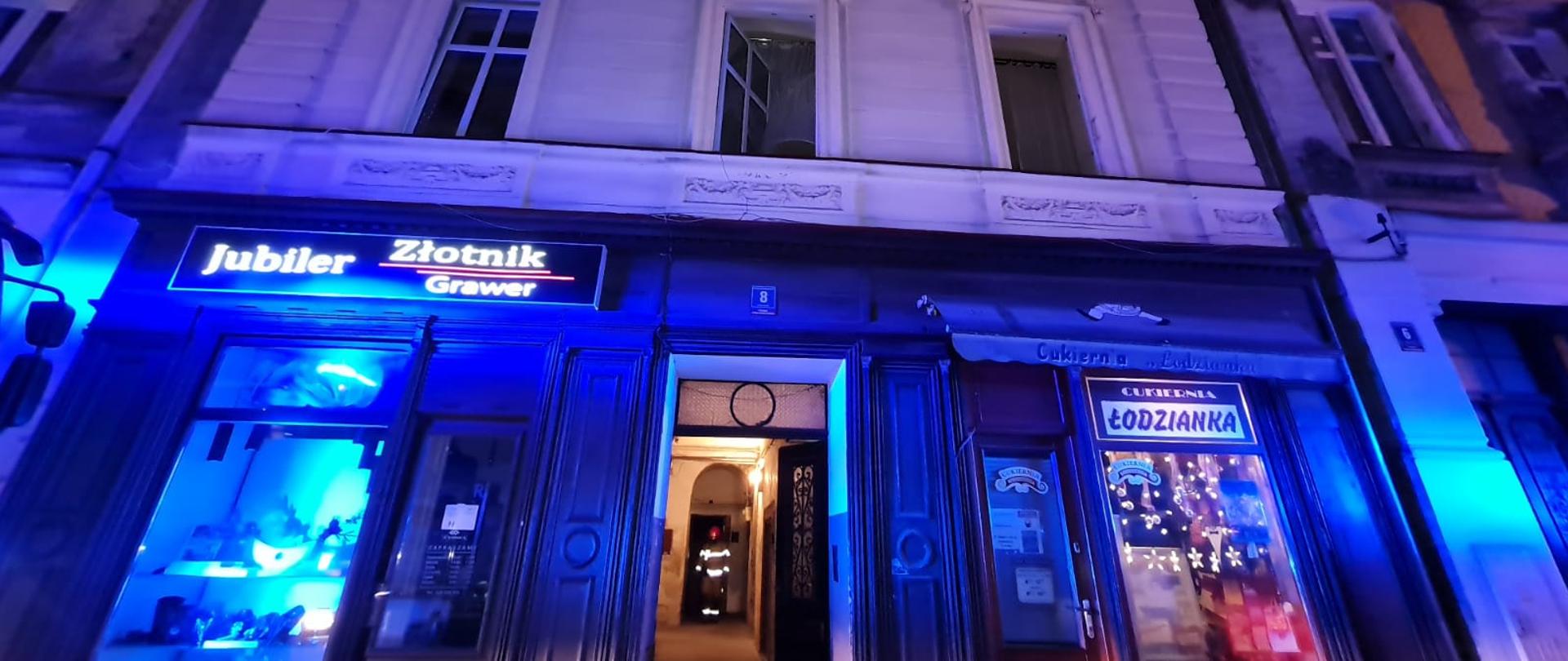 Fasada budynku mieszkalnego w otwartej bramie dostrzegamy sylwetkę strażaka ubranego w ubranie specjalne koloru piaskowego. Na parterze obiektu zlokalizowane dwa lokale handlowe. Okna w budynku otwarte z uwagi na przewietrzanie budynku prowadzone przez jednostki straży pożarnej, Brak widocznego ognia na zdjęciu.