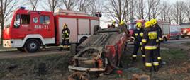 Widoczne miejsce zdarzenia uszkodzony czerwony samochód osobowy oraz służby (strażacy oraz pogotowie ratunkowe działające na miejscu zdarzenia)