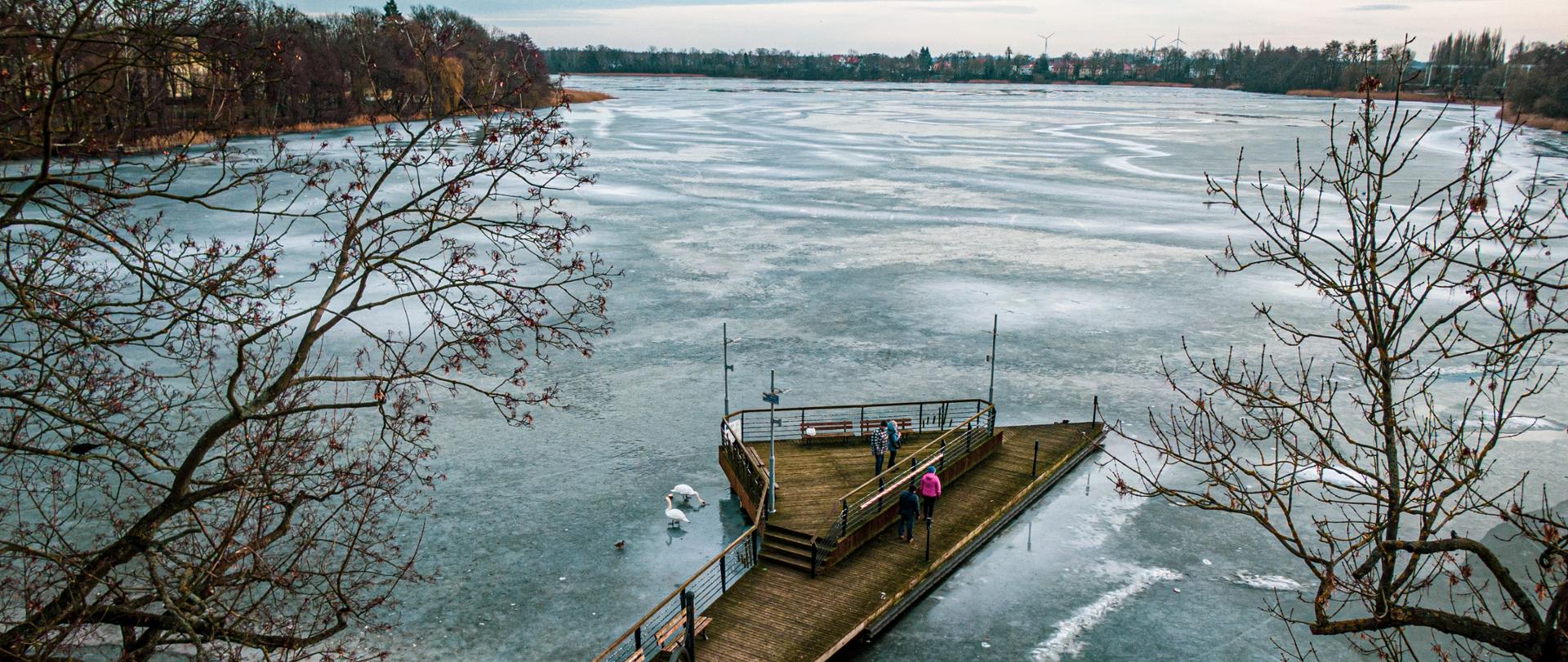 Na zdjęciu znajduje się choszczeńskie jezioro Klukom. Zdjęcie wykonano w dzień przy pochmurnym niebie. Powierzchnie wody pokrywa lód. W dolnej części fotografii znajdują się drzewa, a pomiędzy nimi pomost. Na pomoście znajdują się ludzie, którzy dokarmiają łabędzie.