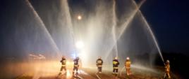 Zdjęcie przedstawia sześciu strażaków trzymających w ręku węże, z których wypuszczana jest w górę woda
Zdarzenie odbywa się wieczorem.