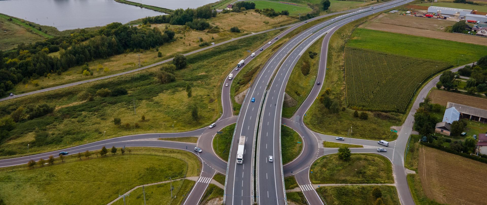 Widok z lotu ptaka na dwujezdniową drogę ekspresową z węzłem drogowym. Jezdnie rozdzielone pasem zieleni, po bokach drogi serwisowe i pola uprawne. W tle widoczne zbiorniki wodne, zabudowa miejscowości oraz rozległy krajobraz rolniczy pod pochmurnym niebem