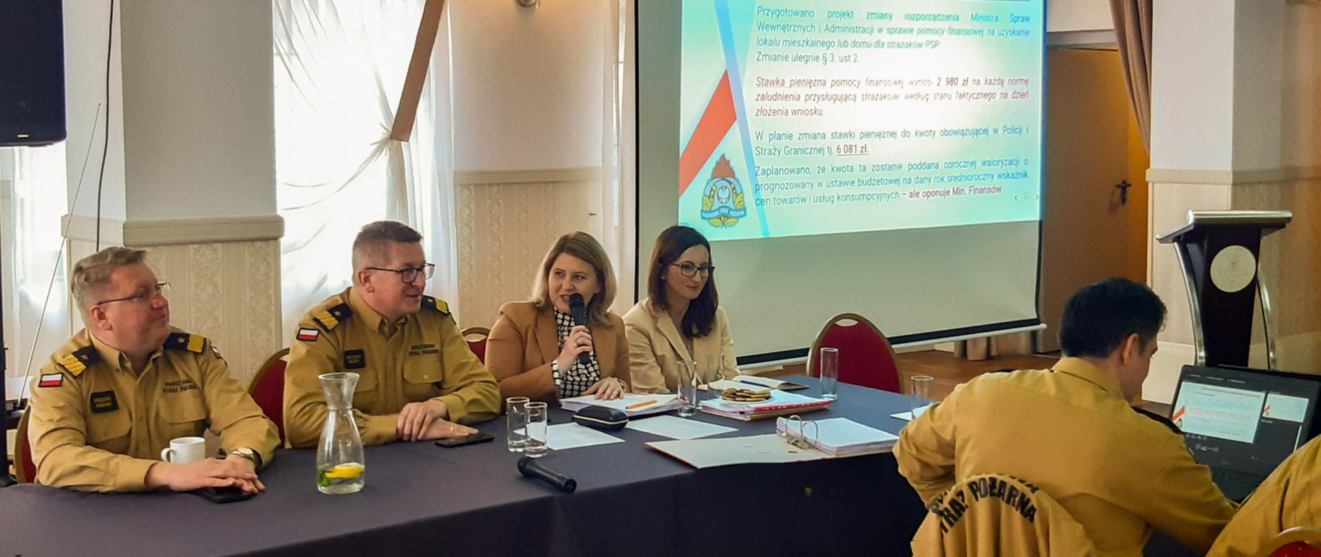 na zdjęciu 3 strażaków PSP oraz dwie kobiety w sali, 4 osoby siedzą przy stole za nimi ekran rzutnika z wyświetlona prezentacją