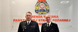Dwóch oficerów PSP w mundurach wyjściowych en face na tle godła Państwowej Straży Pożarnej. Po lewej mł. bryg. Tomasz Samborski, po prawej st. kpt. Marek Wieczorek.