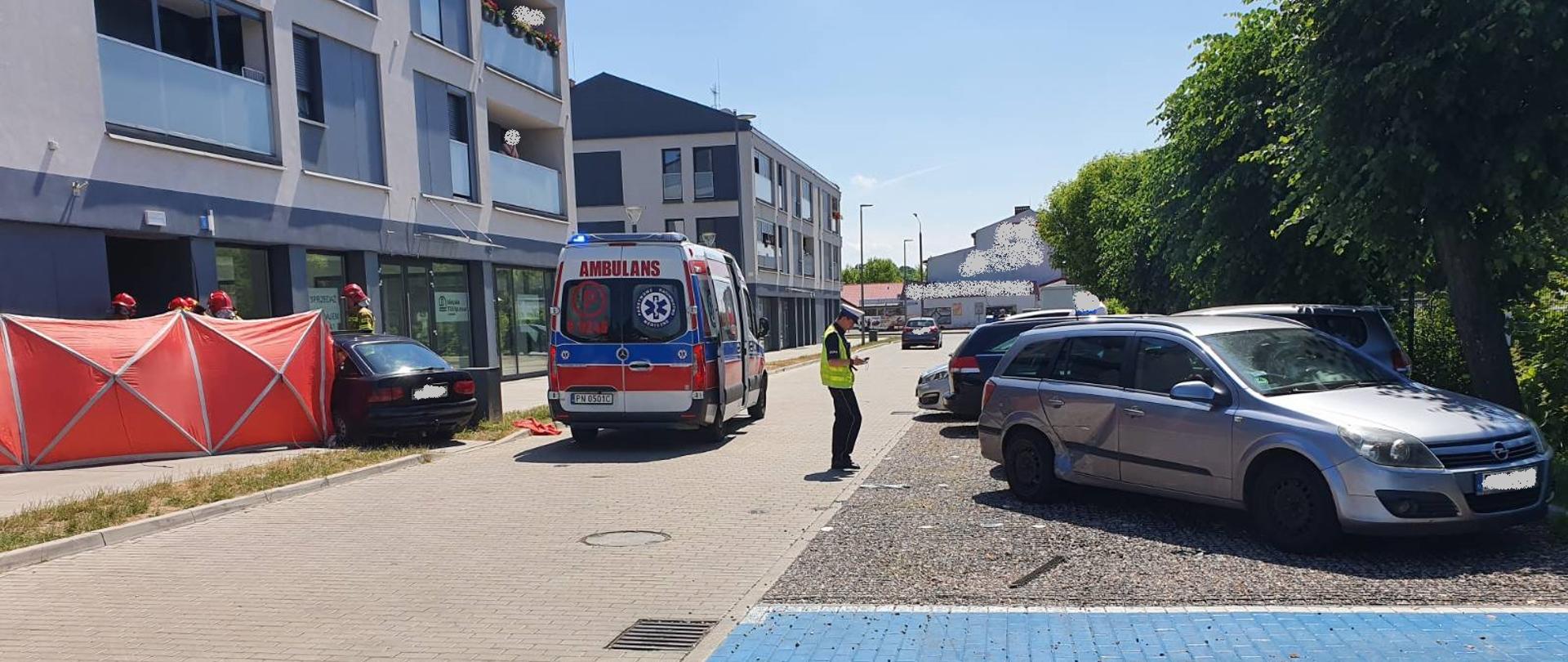 Zdjęcie przedstawia samochód osobowy, który uszkodził dwa zaparkowane samochody, a następnie uderzył w budynek mieszkalny wielorodzinny. Na zdjęciu widać również rozstawiony parawan, karetę pogotowia, strażaków oraz policjanta, 