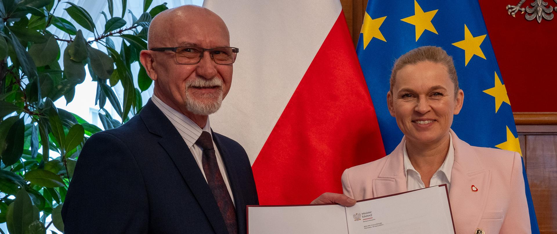 Minister Barbara Nowacka stoi obok mężczyzny w garniturze, nowego dyrektora ORE, Wspólnie trzymają w rękach akt powołania. W tle flagi Polski i UE. 