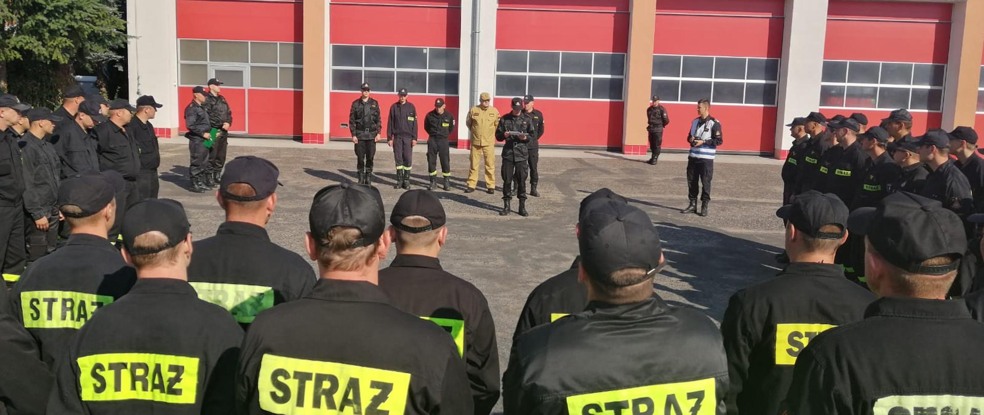 Strażacy stoją na placu komendy. W tle widoczne pomieszczenia garażowe.
