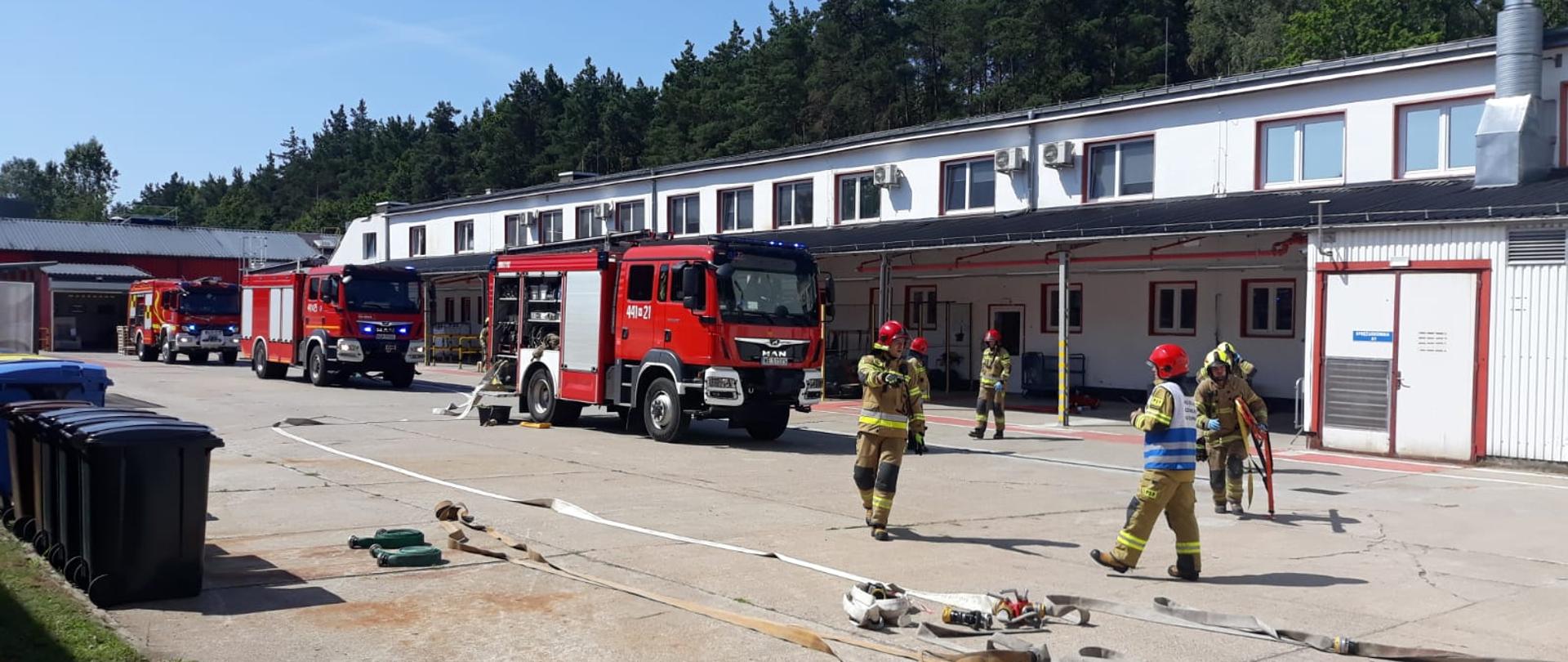 Samochody pożarnicze na terenie firmy 3M, rozwiniete pomiedy nimi węże pożarnicze. Od pierwszego samochodu rozwinięta linia gaśnicza z węży pożarniczych po placu przemieszczają sie strażacy w ubraniach specjalnych.