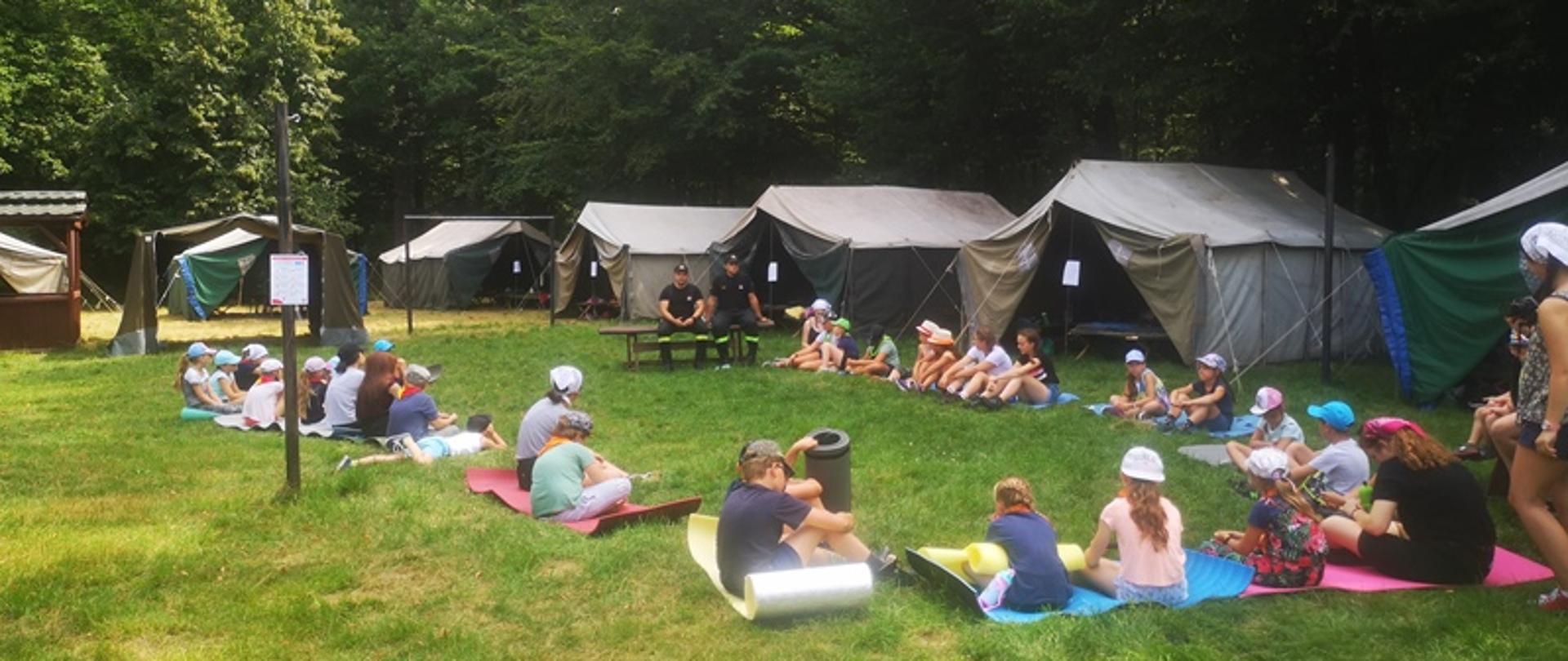 Na ławeczce w pobliżu namiotów siedzi dwóch strażaków w ubraniach koszarowych. Wokół strażaków dzieci uczestniczące w obozie harcerskim wykonały okrąg zasiadając na karimatach ułożonych na trawie. Teren wyznaczony na pogawędkę edukacyjną to prawdopodobnie boisko, ponieważ w tle widać bramkę. Całość terenu otaczają drzewa. Jest słoneczna pogoda. Dzieci ze skupieniem słuchają strażaków, którzy opowiadają o bezpieczeństwie